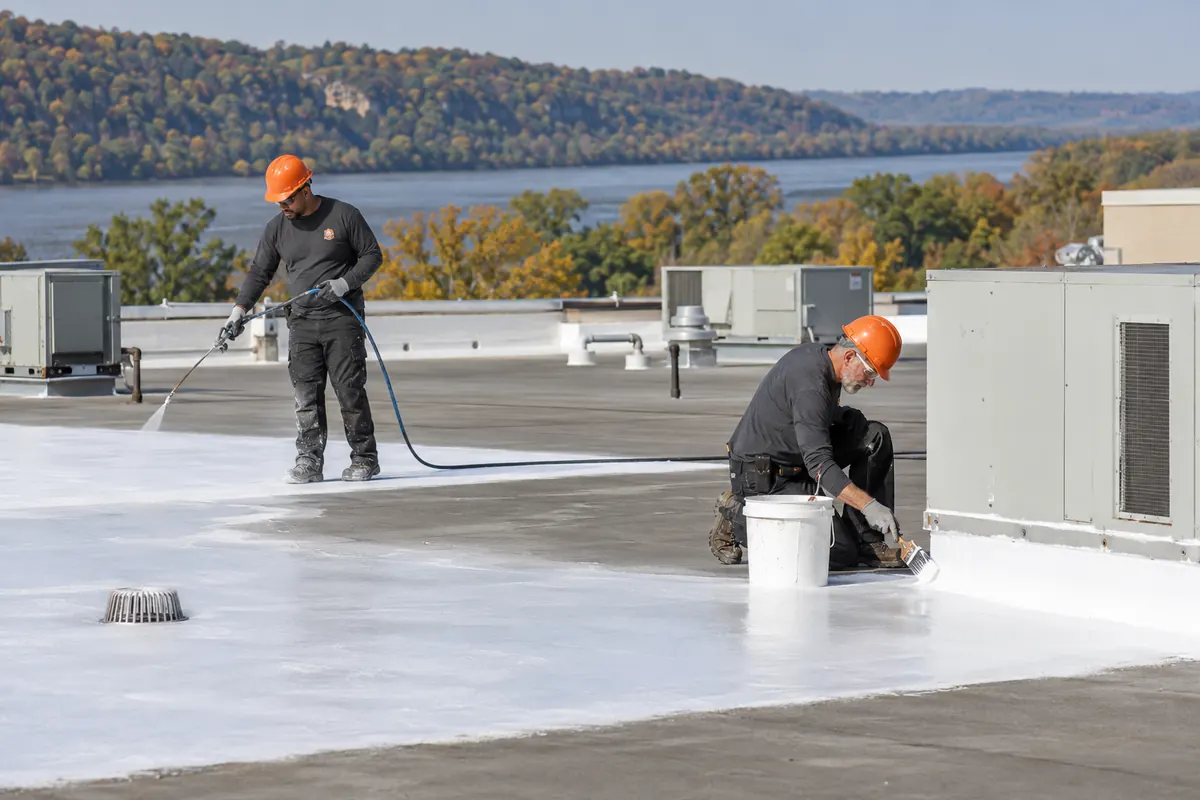 Roofer spraying silicone coating beside rooftop units on a flat commercial roof